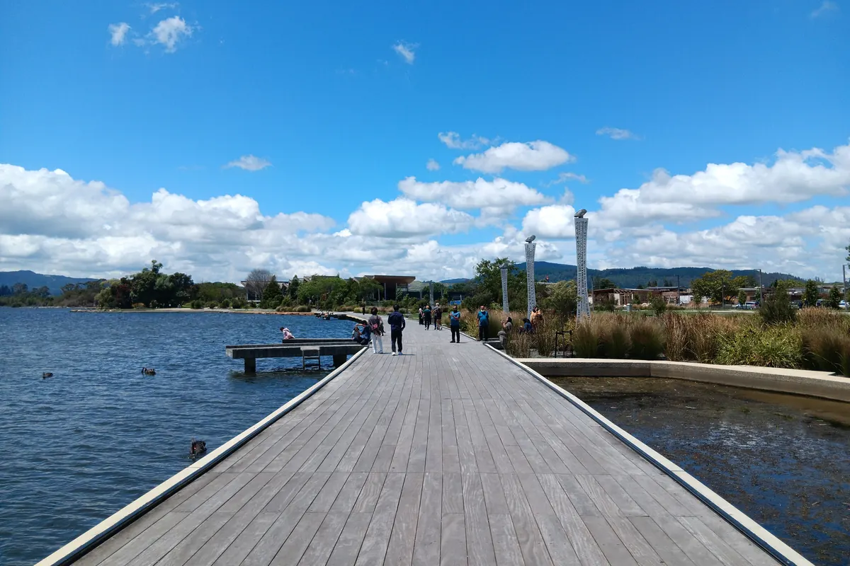 A long timber boardwalk runs out beside the water of Lake Rotorua under a blue sky with broken cloud